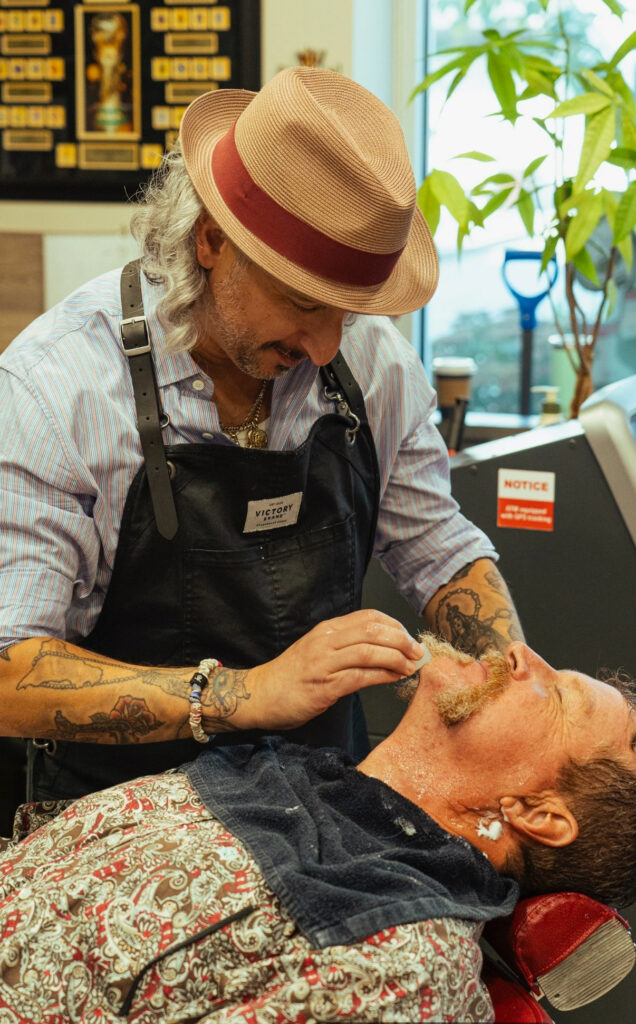 Skilled barber with long gray hair and tattoos wearing a straw hat with red band and black apron, carefully shaving a client's face in Abbotsford barber shop