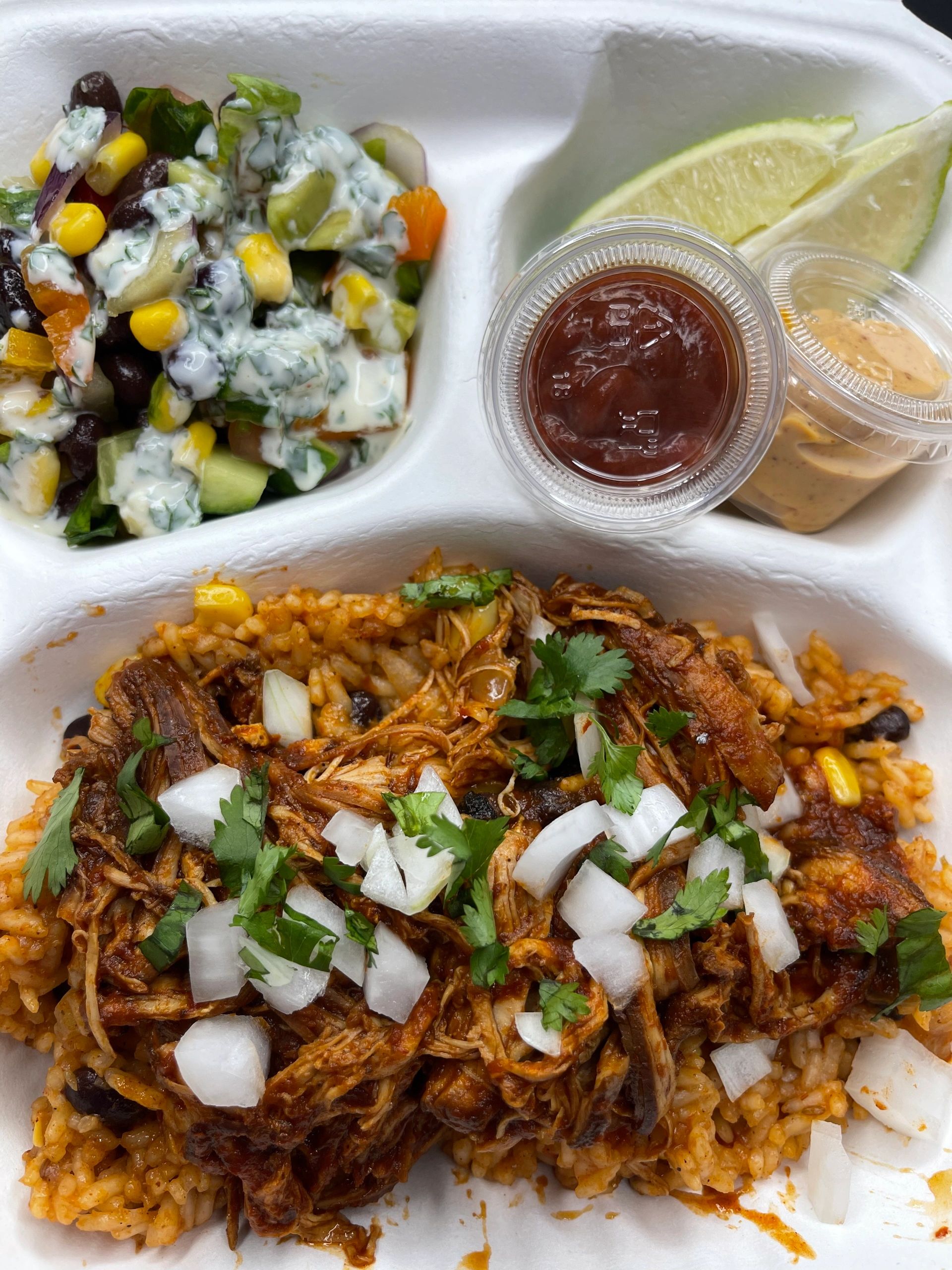 Overhead view of a plated meal featuring shredded birria meat with diced onions and cilantro on Spanish rice, served with a side of creamy vegetable salad with corn and beets, lime wedges, and two sauce containers on a white compartmented plate from a restaurant in Abbotsford