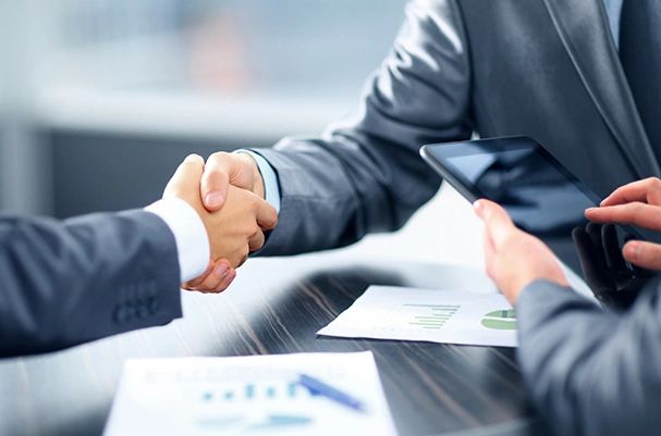 Two business professionals in suits shaking hands during a meeting in Abbotsford, with financial documents and charts visible on the table
