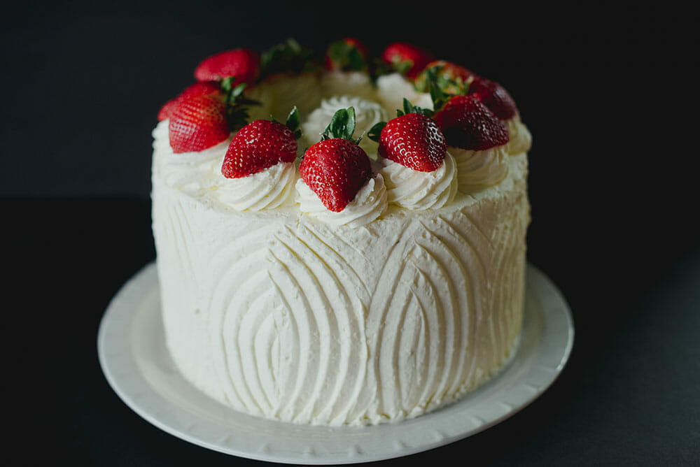 Elegant white frosted cake topped with fresh red strawberries on a white plate against a dark background
