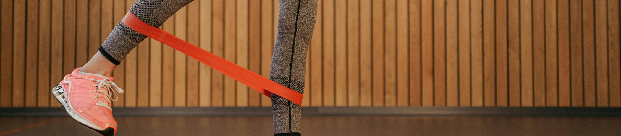 Runner wearing pink shoes and gray leggings jumping over a red resistance band in front of a corrugated wooden wall backdrop in Abbotsford