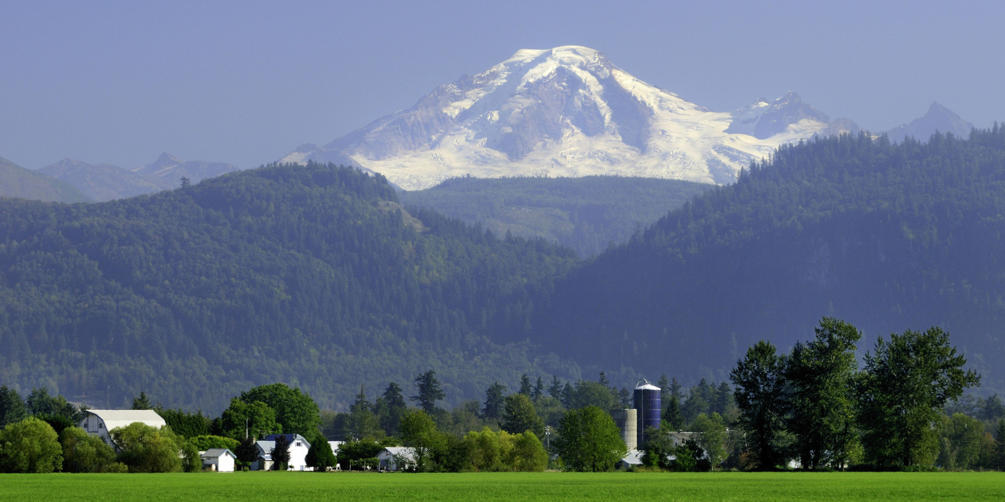 Mount Baker snow-capped peak rises above forested mountains with farmland and rural buildings in Abbotsford valley in the foreground