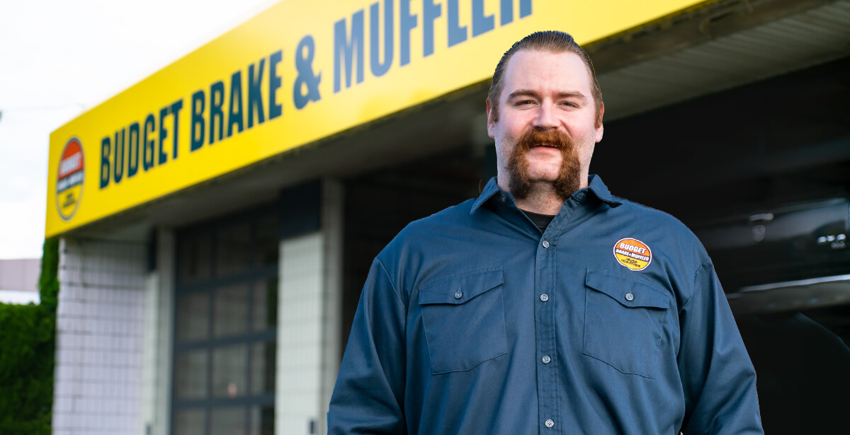 Sam stands in front of Budget Brake & Muffler shop in Abbotsford wearing a blue work shirt with company logo