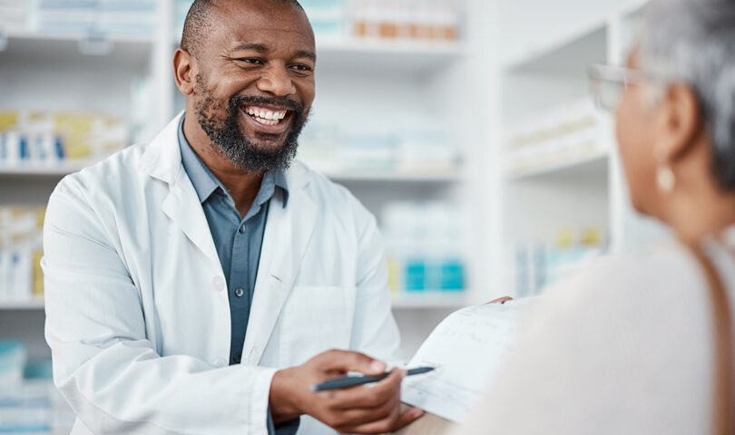 Friendly male pharmacist in white coat and blue shirt smiling while assisting a female customer at the Pharmasave Clayburn Centre pharmacy in Abbotsford, with medication shelves visible in the background
