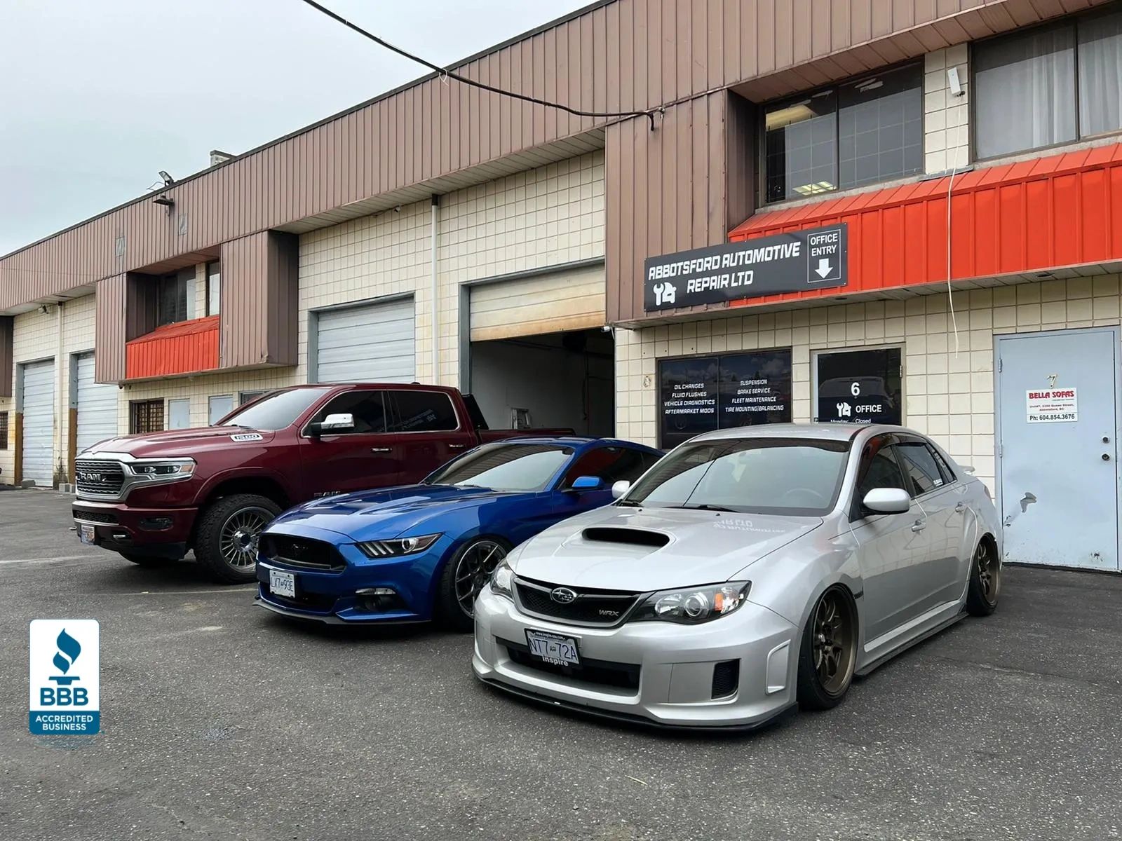Three performance cars parked outside Abbotsford Automotive Repair Ltd facility - a burgundy RAM truck, blue Ford Mustang, and silver Subaru WRX