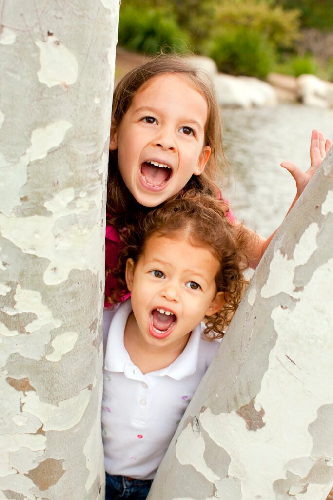 Two happy young children peeking out from behind a white textured tree trunk outdoors, smiling joyfully at the camera in Abbotsford