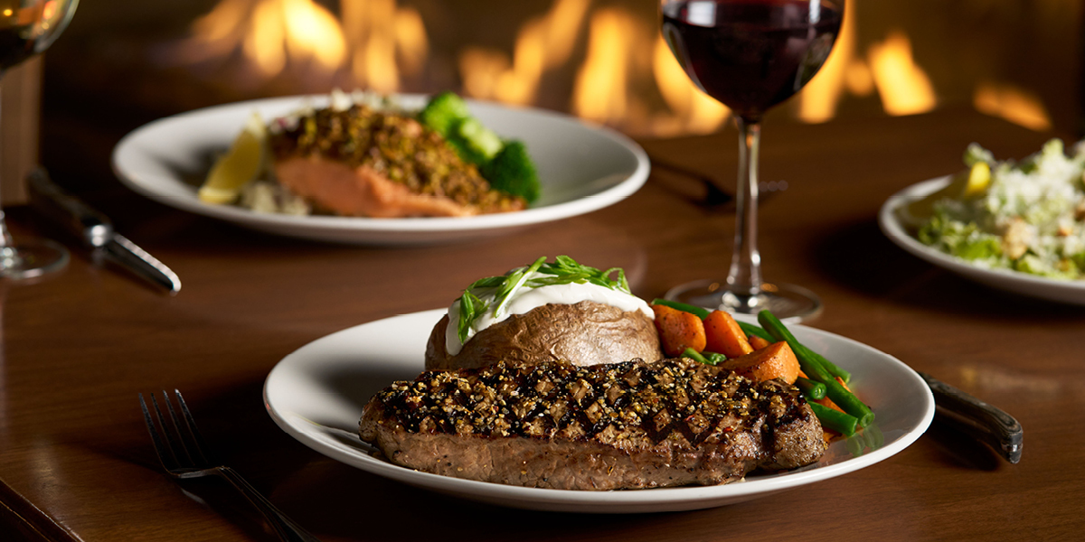 Gourmet steak dinner with sesame-crusted meat, roasted vegetables, and baked potato topped with sour cream and chives, served at The Keg Steakhouse + Bar in Abbotsford, accompanied by red wine