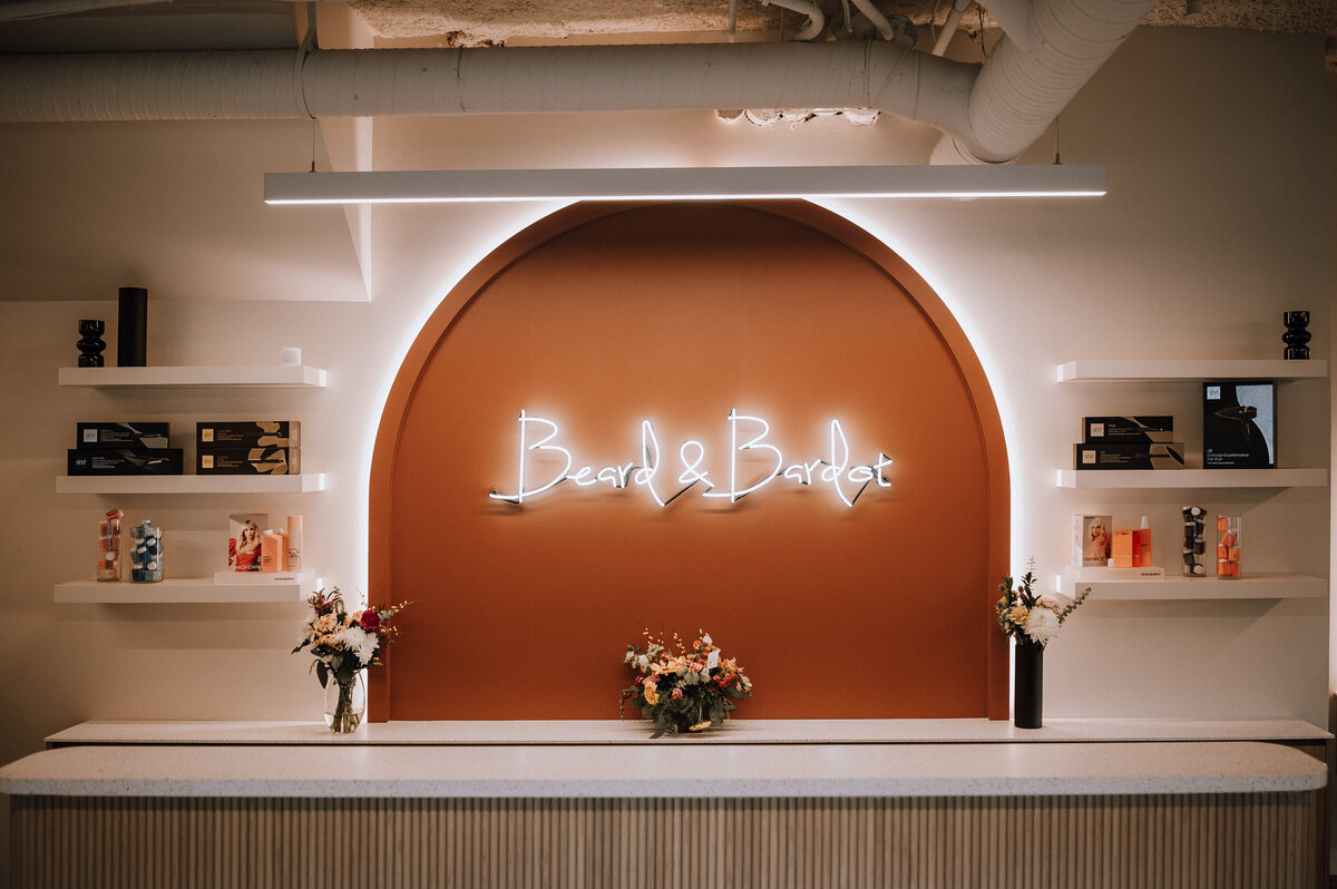 Beard & Bardot Hair + Skin Co. salon interior in Abbotsford featuring a modern reception desk with white neon signage on an orange curved accent wall, flanked by product shelves and fresh flowers