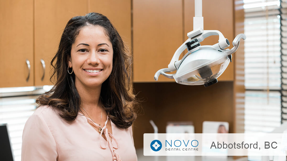 Dr. Provo smiling in her dental operatory at NOVO Dental Centre in Abbotsford, BC, with overhead dental light and wood cabinetry visible in the background