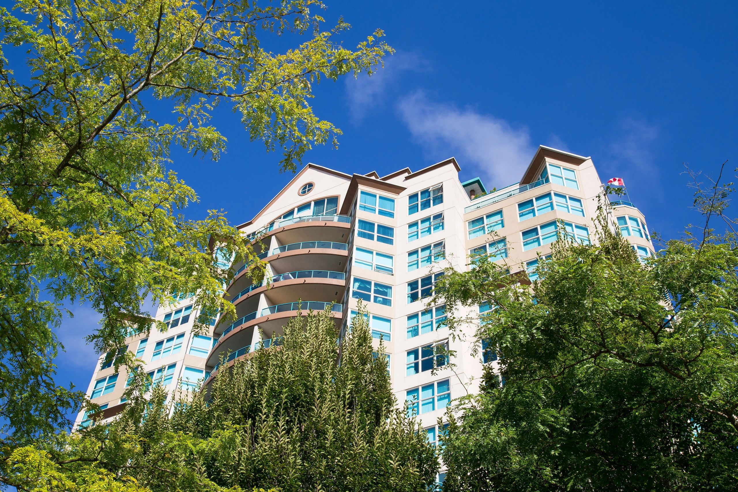 Garden Park Tower in Abbotsford, a multi-story residential building with cream and white facade featuring curved balconies and blue-tinted windows, surrounded by green trees against a clear blue sky