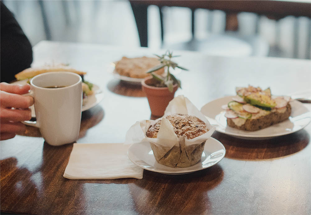A cozy brunch scene at The Polly Fox in Abbotsford featuring a cream-colored mug of coffee, a freshly baked muffin in parchment paper, artfully plated toast with toppings, and potted herbs on a warm wooden table