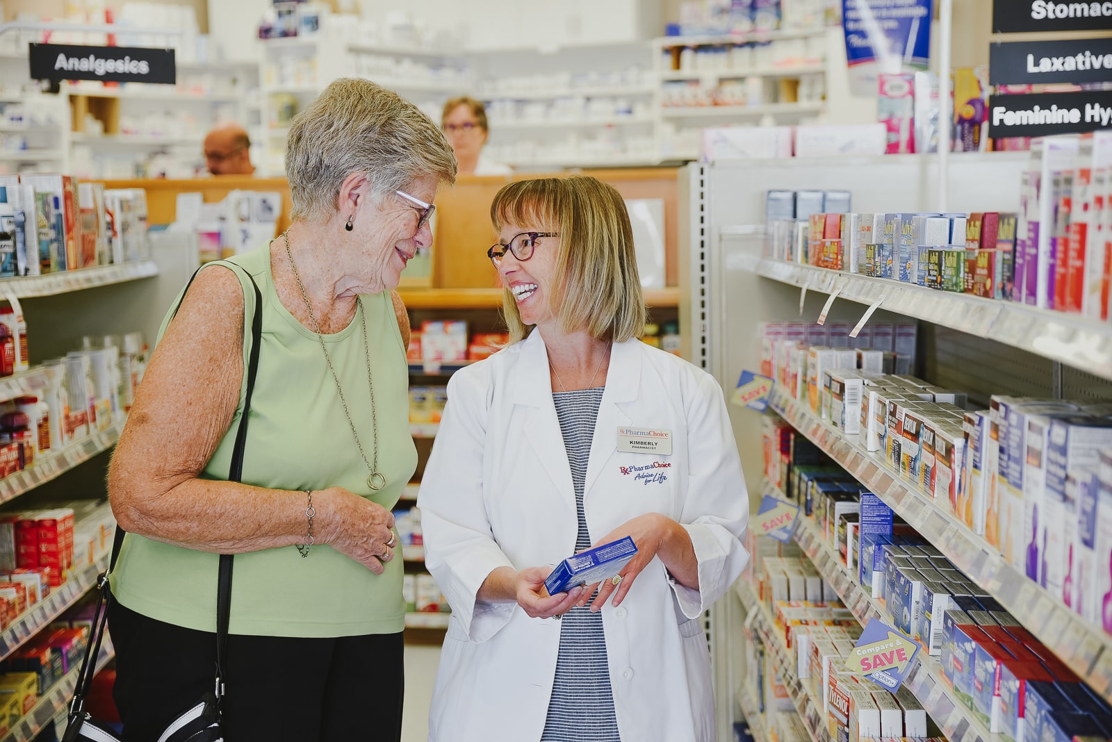 A pharmacist in a white coat assisting a senior customer in the pharmacy section at Versa PharmaChoice Abbotsford, surrounded by shelves of medications and health products.