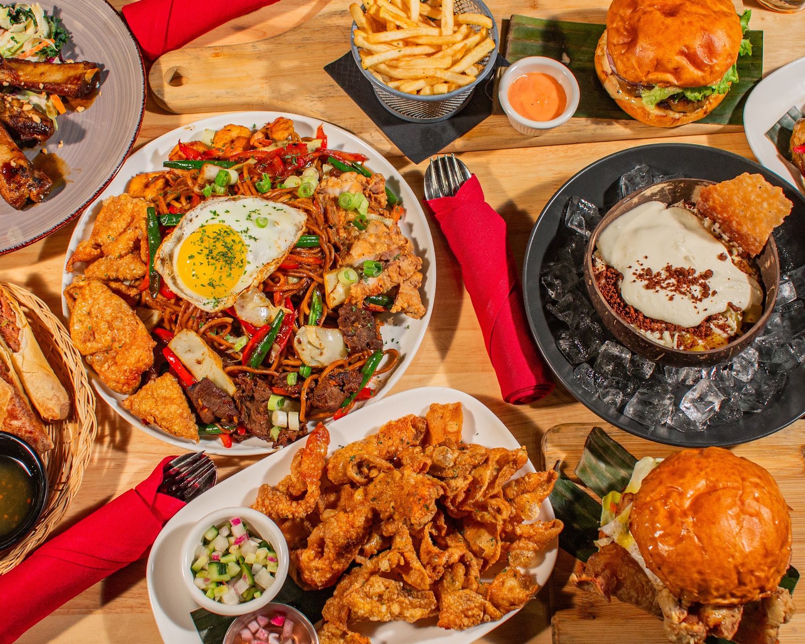 Overhead view of Halina dishes on a wooden table, including Pancit Batil Patong topped with a fried egg, crispy chicken skin, burgers with fries and a dessert set on ice